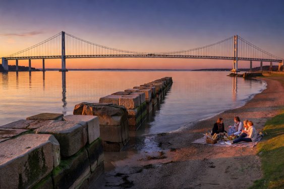Stone piers and waterfront shoreline looking toward an architectural suspension bridge at sunset